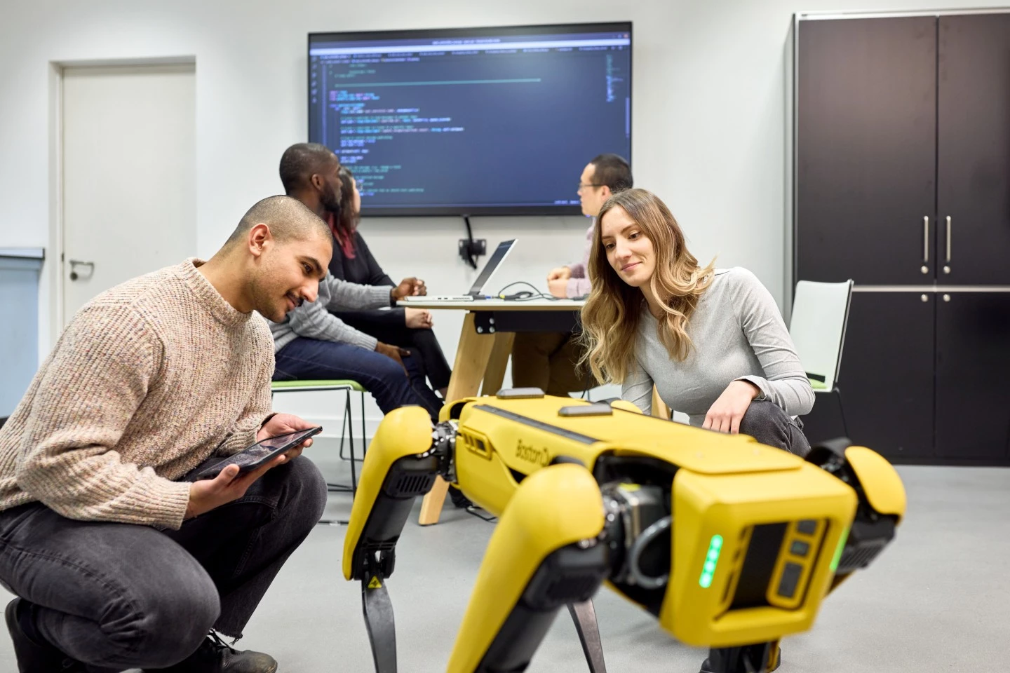 Two students using the Boston dynamics robot while another group of students work on code at a desk