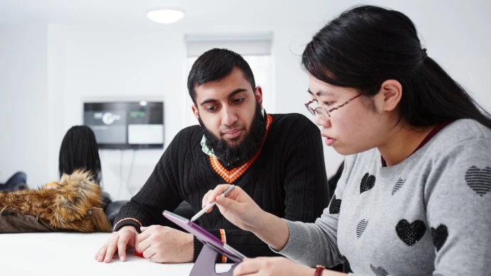 Two students sat in a halls of residence looking at a tablet. 