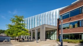 A sleek, modern Business School building made of glass and concrete stands next to a red-brick university building, both surrounded by greenery under a bright blue sky