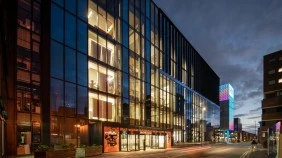 Modern university building with glass facade, illuminated at dawn, surrounded by city buildings