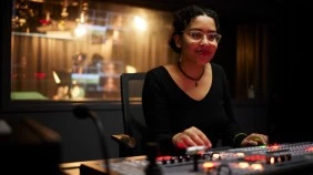 Student in a TV studio control room, with a backdrop of the main studio setup, adjusting levers on the mixing console.