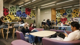 Groups of students sitting on benches and high chairs in a university common space. Some are engaged in work, while others are on their phones, with colourful painted walls as a backdrop.