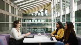 Group of students sitting in study space while engaged in conversation, with a backdrop of a modern Business School building and glass facades