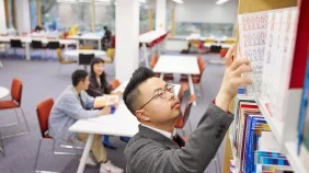 International student in library in the foreground, reaching for a book in book shelf, with a group of students engaging in conversation in the background