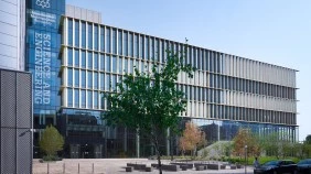 A modern, innovative Science and Engineering university building, surrounded by trees and green space, as well as benches placed outside building. Few cars parked in front and students walking by.