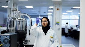 A student in a biology laboratory, wearing safety goggles and a lab coat, is conducting an experiment at a lab bench. The backdrop features another student engaged in experiments, surrounded by lab equipment, creating a dynamic and focused scientific environment.