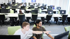 Two students engaged in conversation in a computer lab, surrounded by rows of desktop PCs, desks, and office chairs. One student is pointing at the screen, while the other listens attentively, both focused with a backdrop of fellow students working.