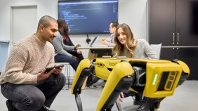 Two students examining and programming innovative robot, with two students engaged in conversation in the background as well as a large screen displaying code, creating a tech-savvy environment