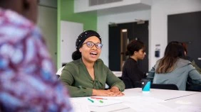 Student smiling and engaged in conversation in a busy seminar room, with a desk of pens and paper in front, surrounded by fellow students.