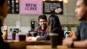 Two international students chatting in café, surrounded by students engaged in conversation, with the café counter and signage in the background