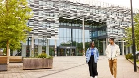 Two students walking along a path outside a modern university building with a glass facade, surrounded by benches and trees