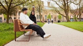 Two students sitting on bench in All Saints Park, surrounded by greenery and trees, with two students walking along paved path in the background