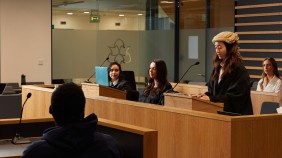 A group of students engaged in a Law School moot court session, with one student standing and reading out a statement while the others attentively listen. The backdrop features large windows and a glass door.