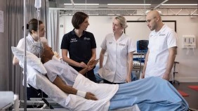 Group of nursing students and a lecturer in a simulation room, standing in front of a hospital bed with a simulated patient.