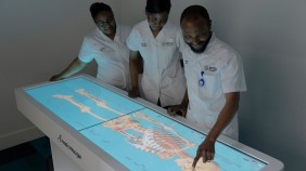 Group of three students in nursing uniforms standing by an Anatomage table, with a lit-up screen displaying a skeleton