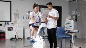 Two students in a sports lab environment: one riding a stationary bike while the other reads from a document, surrounded by sporting equipment